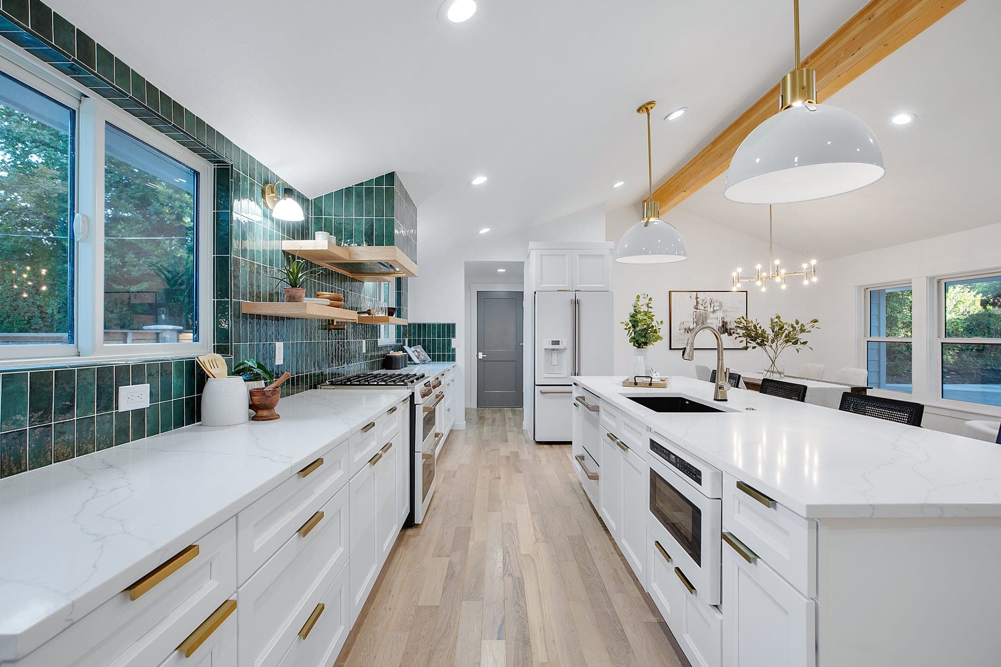 Full kitchen remodel with a green vertical stacked tile backsplash, white shaker cabinetry with gold hardware, quartz counters, a long island with a prep sink, open white oak shelving, and hardwood flooring by Portland Tile in Portland, OR