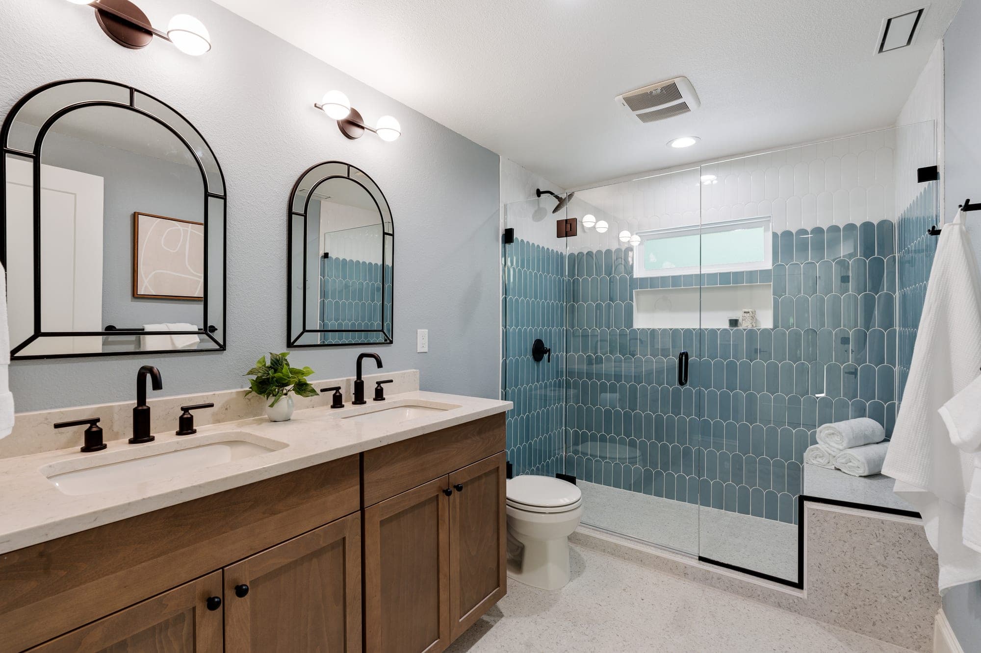 Craftsman primary bathroom with a stained wood double vanity, arched black framed mirrors, a blue and white fish scale tile shower, a terrazzo floor and bench, and frameless glass in downtown Portland, OR