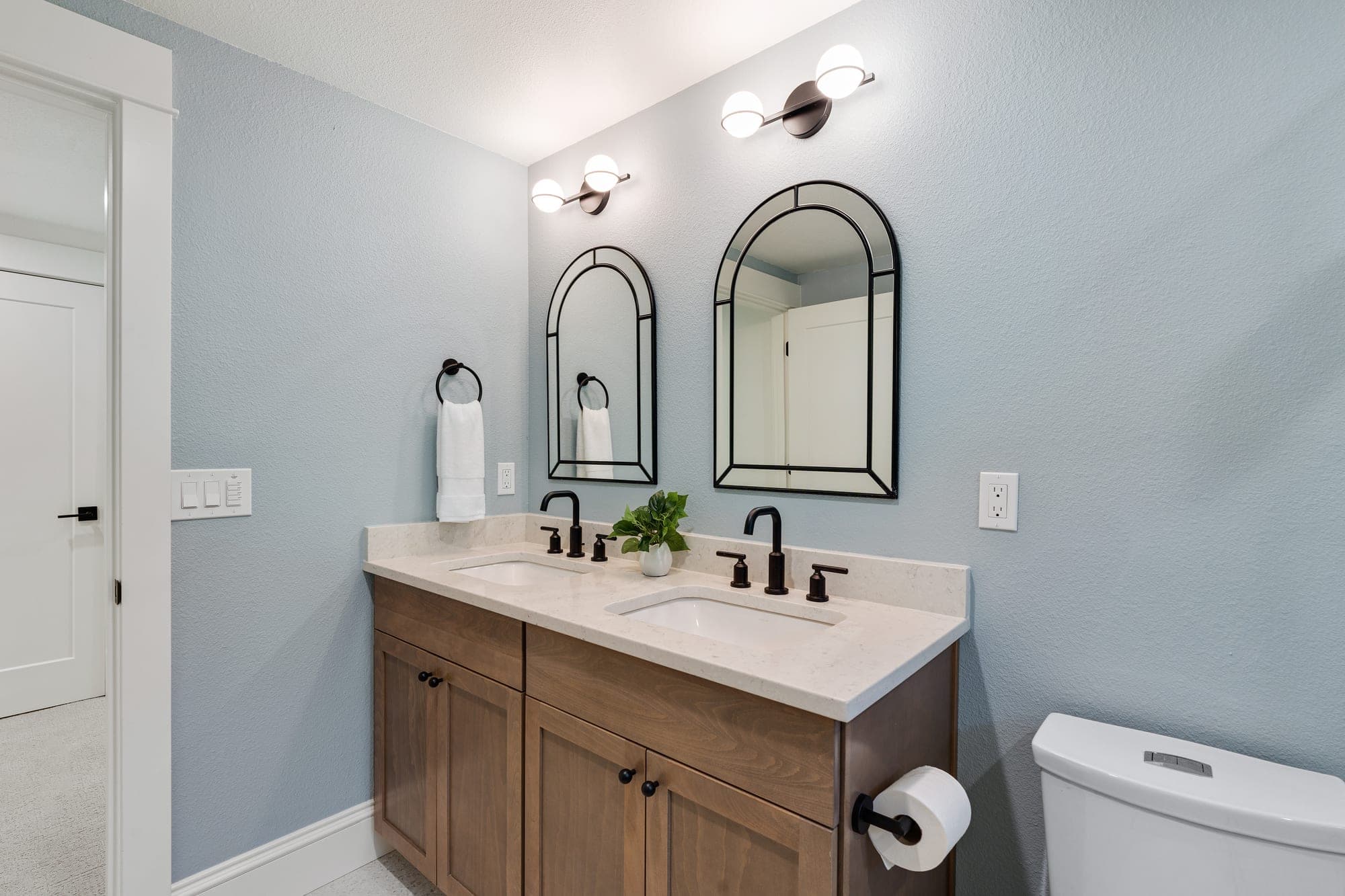 Stained wood double sink vanity with arched black framed mirrors, black faucets, a quartz counter, and pale blue walls in a craftsman bathroom in downtown Portland, OR