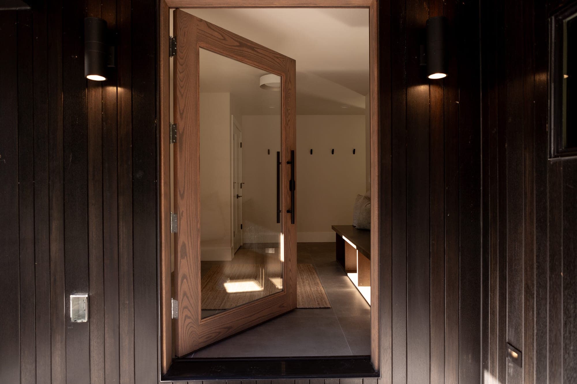 Entry with a large format tile floor seen through a wood framed glass pivot door, with a built in bench and dark vertical siding by Portland Tile in Portland, OR