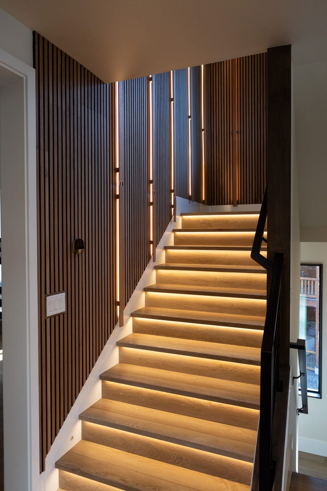 Modern staircase with lit treads, a vertical wood slat wall, and a black metal railing by Portland Tile & Remodels