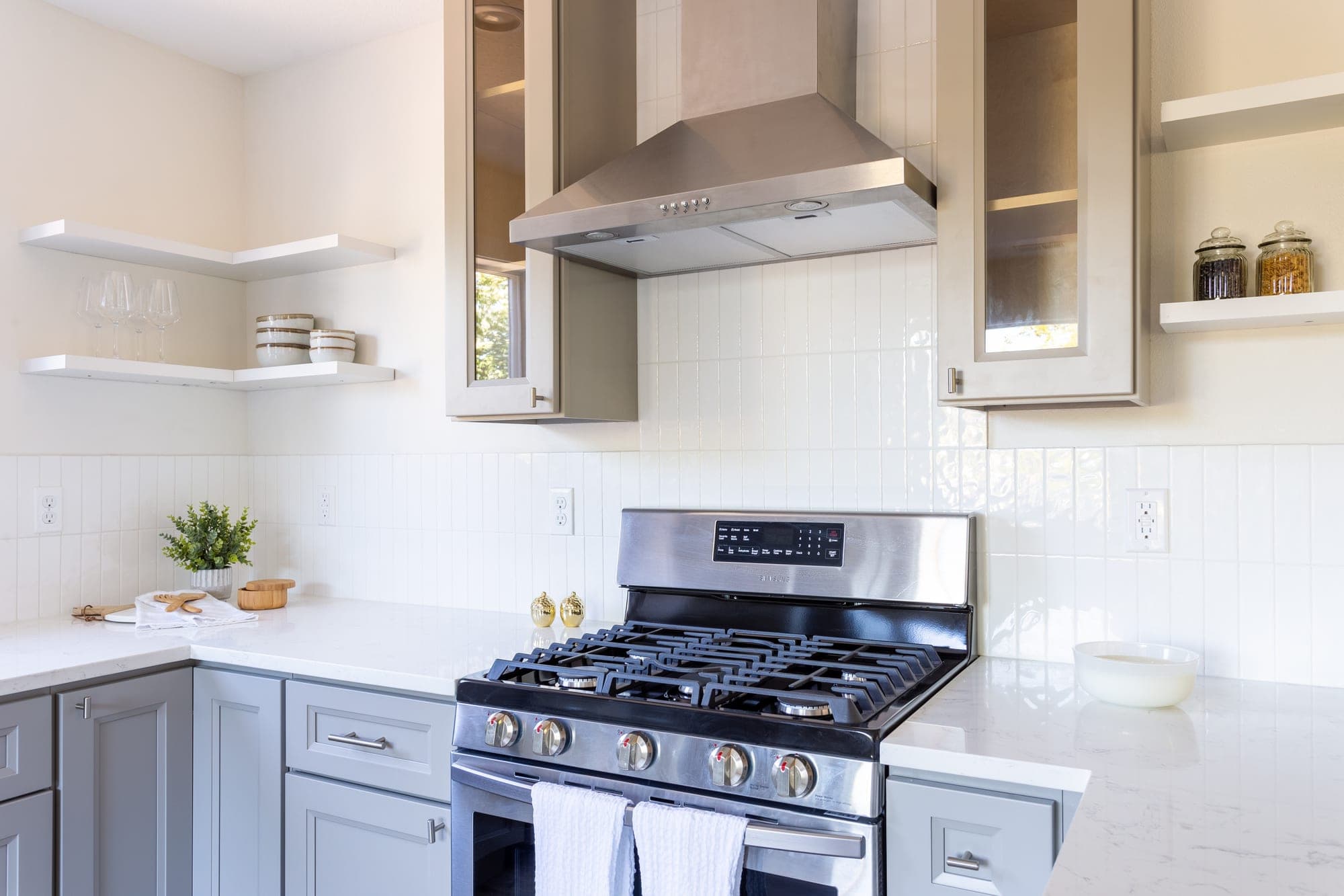 Stand alone stainless steel range hood flanked by glass front cabinets above a gas range with a white vertical stacked tile backsplash in Hillsboro, OR