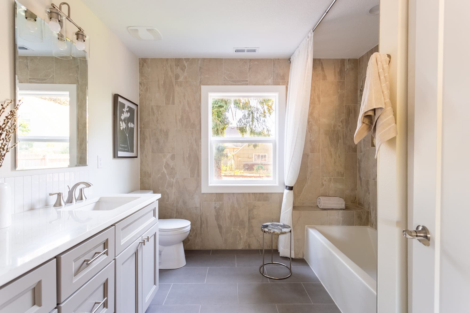 Remodeled primary bathroom with a tile feature wall wrapping the window, a tub and shower in large format tile, a white vanity with a quartz counter, and gray tile flooring in Hillsboro, OR