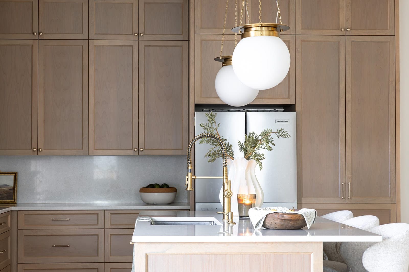 White oak kitchen island with the sink set into it, a gold spring faucet, gold globe pendants, quartz counter, and a paneled stainless steel refrigerator behind in Camas, WA